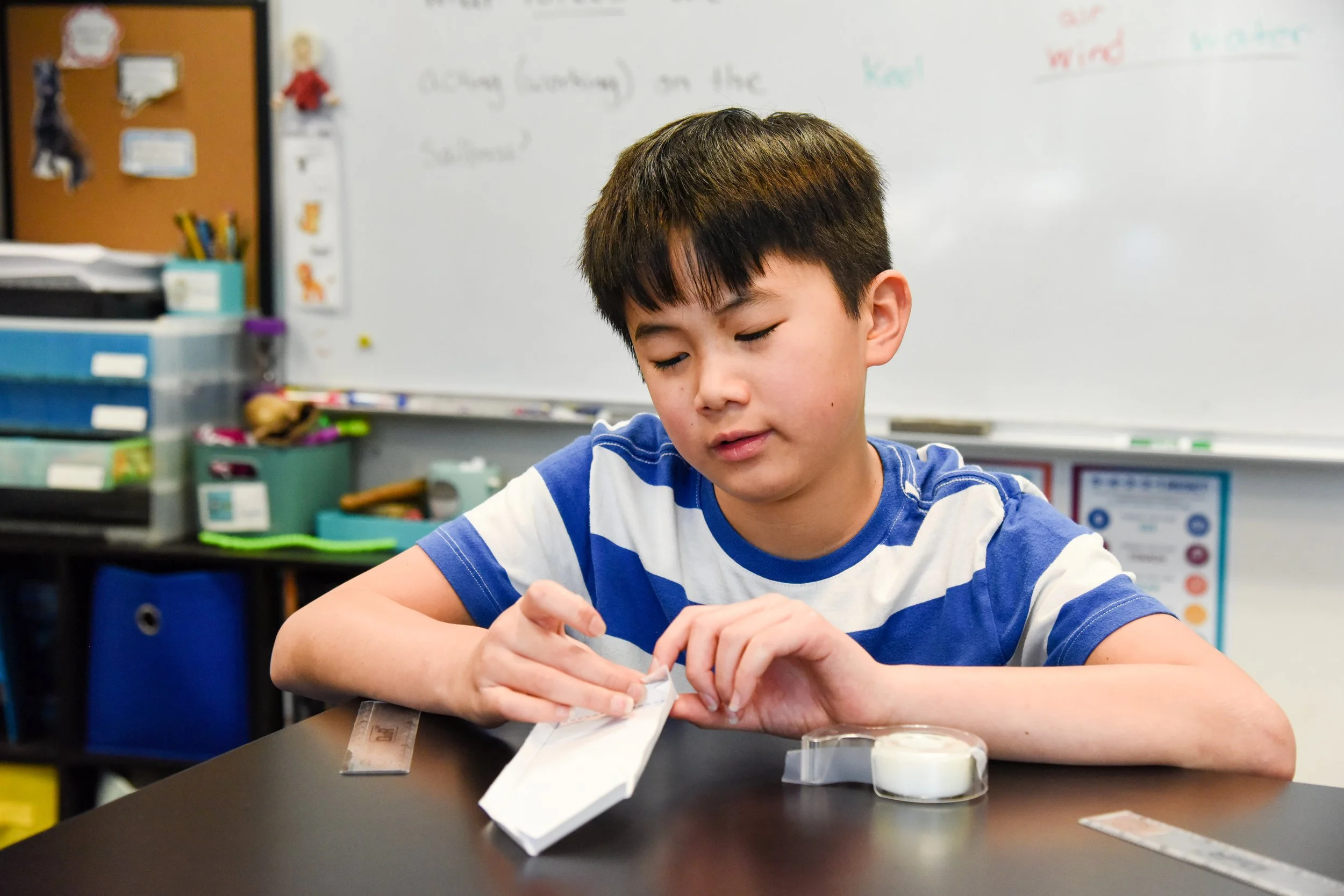 A young boy with dark hair and wearing a blue and white striped shirt is sitting at a school desk, focusing on a project. The classroom background includes a whiteboard with colorful writing and various storage containers on shelves.