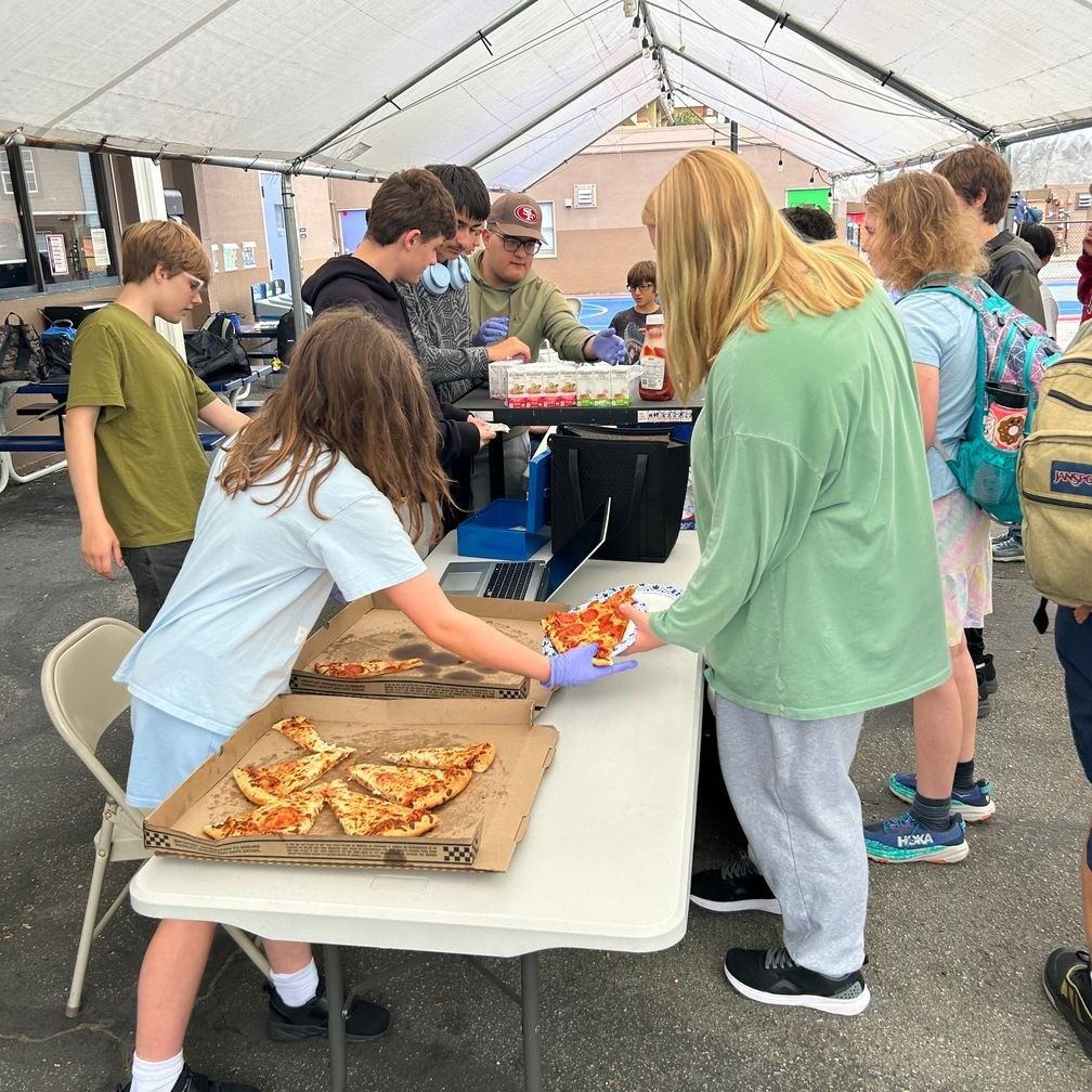 A group of people, including children and adults, are gathered around a table under a tent, eating pizza and receiving drinks at an outdoor event.