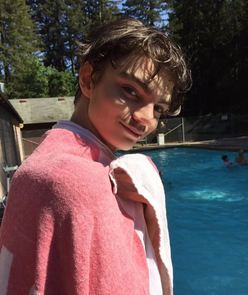 A young person with wet hair smiling at the camera, standing near a swimming pool on a sunny day, with trees in the background.