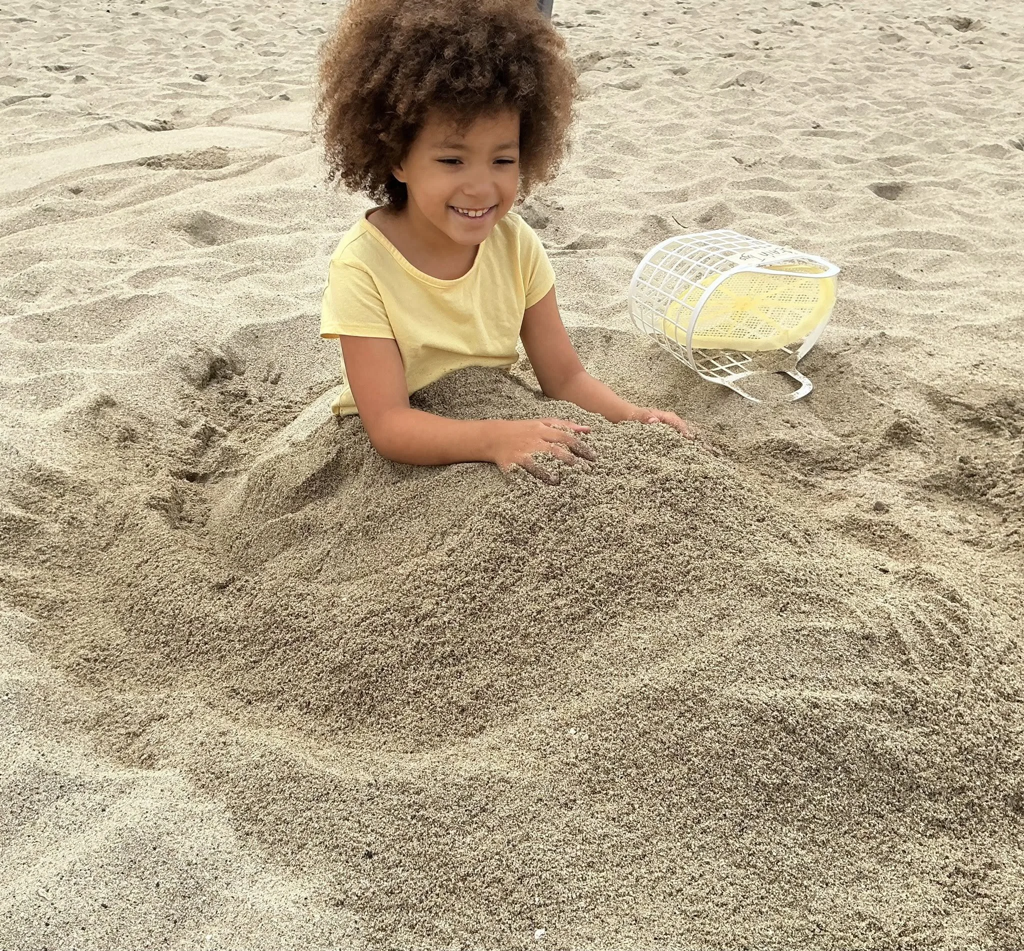 A young girl with curly brown hair wearing a yellow shirt sitting in the sand on a beach, smiling, with a white plastic brush nearby.