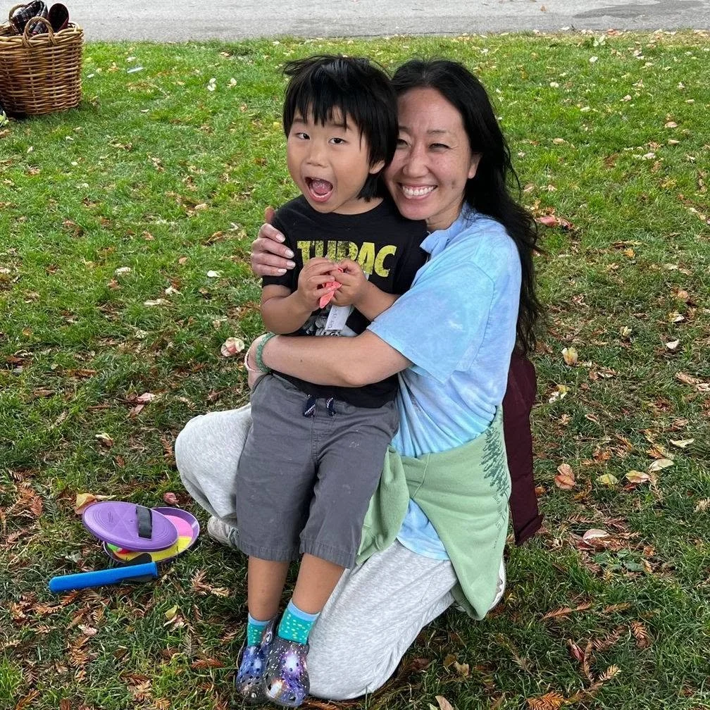A woman kneeling on grass in a park, hugging a young boy. Both are smiling and happy. There are scattered leaves on the ground, and a basket with items and a toy in the background.