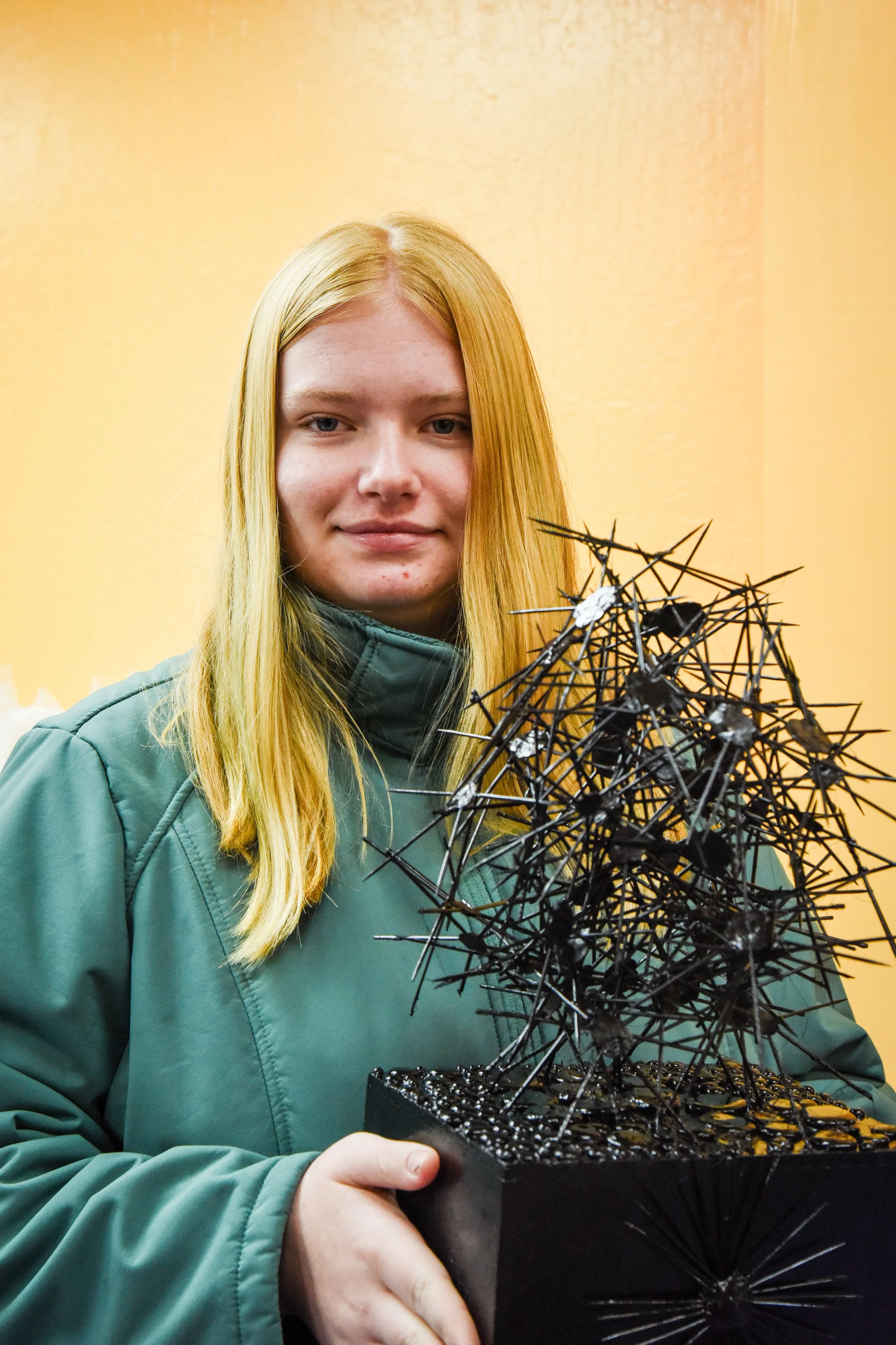 A young woman with long blonde hair holding a black geometric art sculpture against a light yellow wall.