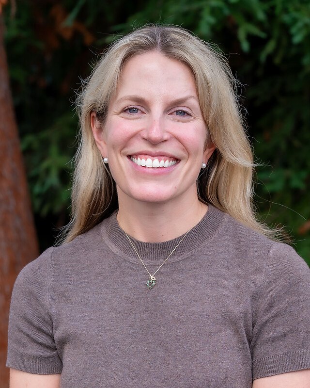 A woman with long blonde hair smiling outdoors, wearing a gray top and a necklace with a floral pendant, with greenery in the background.