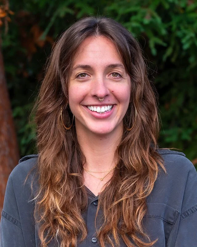 A woman with long, wavy brown hair, wearing earrings, a septum piercing, a necklace, and a gray shirt, smiling outdoors with green foliage in the background.