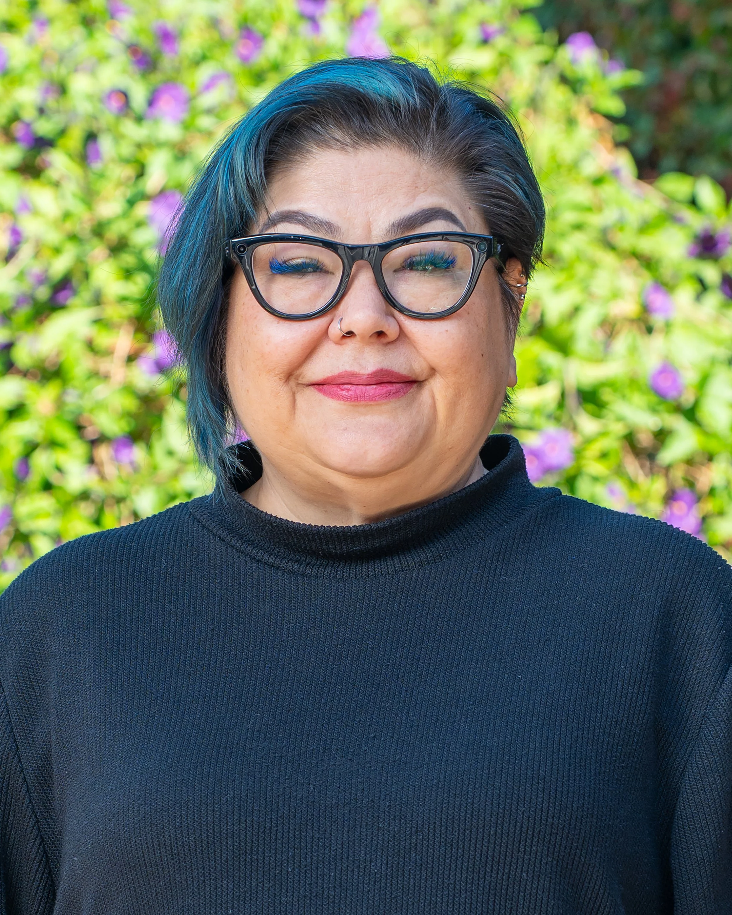 A woman with short black hair with blue streaks, wearing black glasses and a nose piercing, standing outdoors with green foliage and purple flowers in the background.