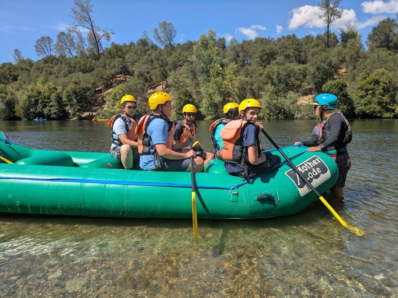 Group of people in a green inflatable raft on a river, wearing yellow helmets and life jackets, with a guide in a blue helmet in the water.