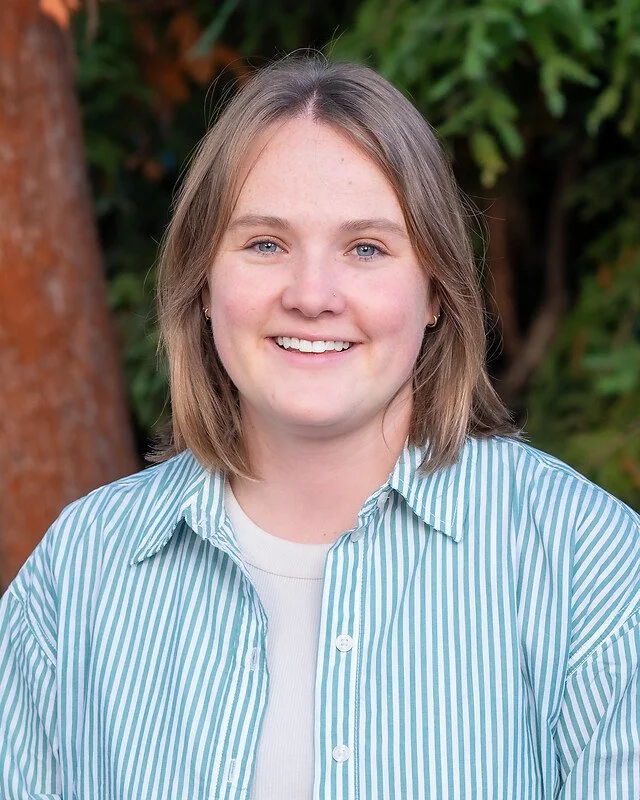 A young woman with shoulder-length light brown hair, blue eyes, and a nose piercing, smiling outdoors in front of green foliage and a tree.”}