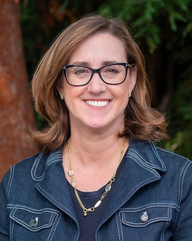 A woman with light skin, shoulder-length brown hair, and glasses smiling outdoors, wearing a navy denim jacket and jewelry.
