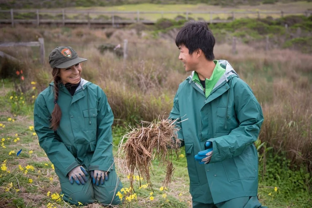 Two people in rain gear, a woman with a braid and a man with short hair, are outdoors in a grassy area, smiling at each other while holding a clump of grass roots.