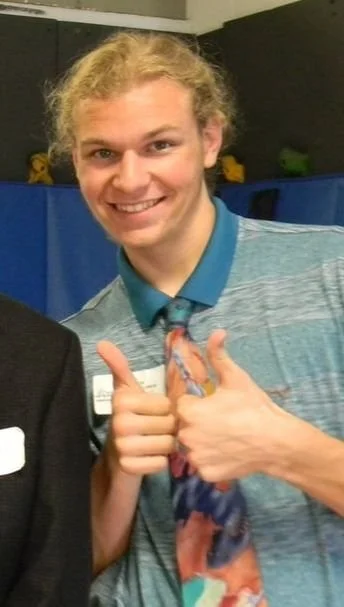 Young man with curly blond hair smiling, wearing a blue polo shirt, giving a thumbs-up gesture.