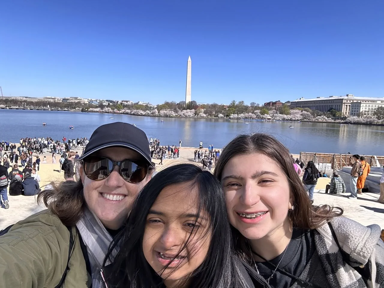 Three women taking a selfie in front of the Washington Monument across the Reflecting Pool in Washington, D.C., with cherry blossoms and a crowd of people in the background.