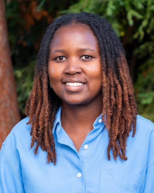 Young woman with shoulder-length dreadlocks wearing a blue button-up shirt, outdoors with green foliage in background.