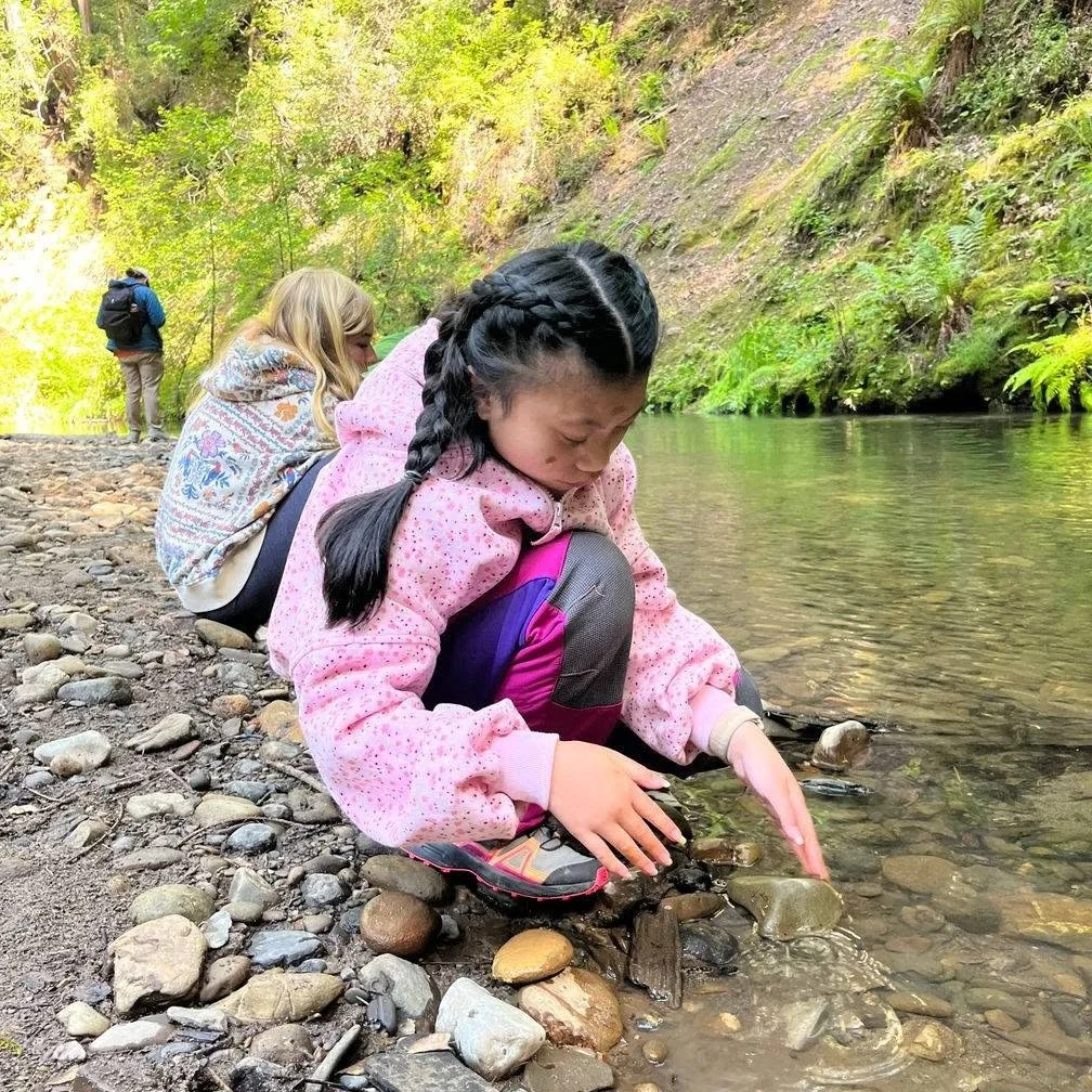 A young girl crouches by a river's edge, kneeling on rocky ground, with her hands reaching into the water. She has black hair styled in a braid and wears a pink fleece jacket and colorful pants. In the background, a woman with long blonde hair, wearing a patterned shawl, and a man with a backpack and cap are also near the water, surrounded by lush green trees and a rocky hillside.