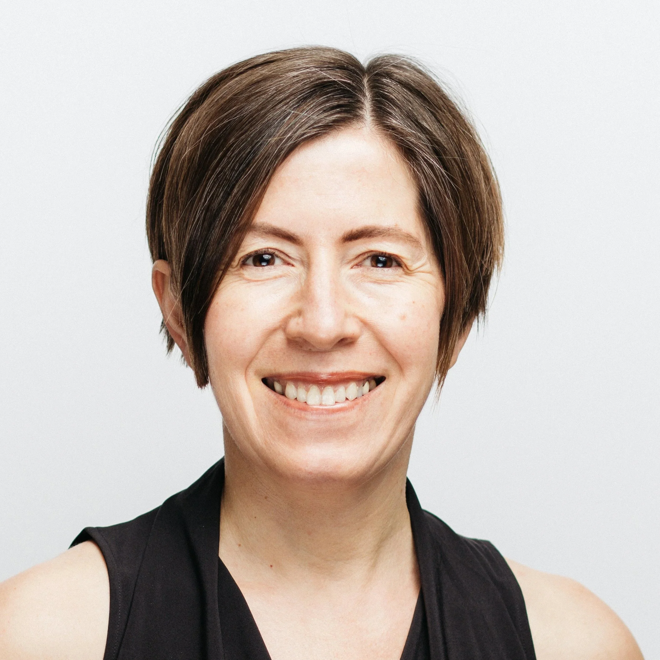 Close-up of a woman with short brown hair smiling, wearing a black sleeveless top, against a light gray background.