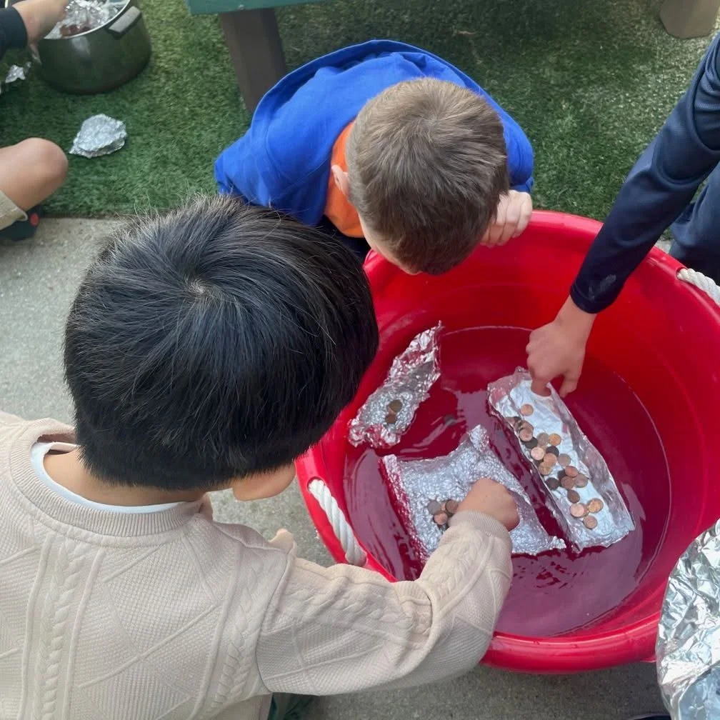 Children collecting coins from aluminum foil in a red water tub during an outdoor activity.