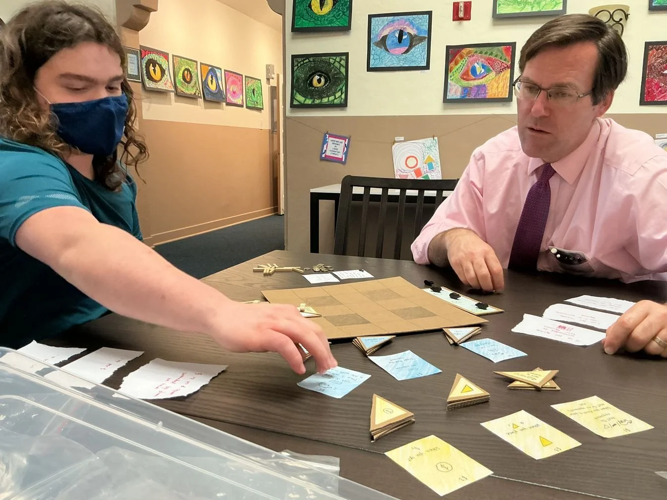 Two people sitting at a table playing a game with triangular tiles. The student the left is wearing a blue mask and teal shirt, and the teacher on the right is wearing glasses, a light pink shirt, and a purple tie.