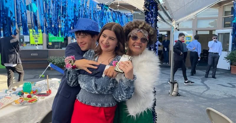Three people smiling and hugging at an outdoor school dance, with blue and green decorations hanging above and other people in the background.