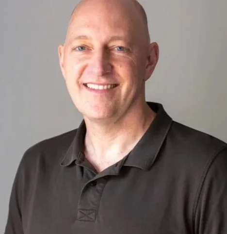 Bald man with blue eyes and a big smile, wearing a black polo shirt, standing against a light gray background.
