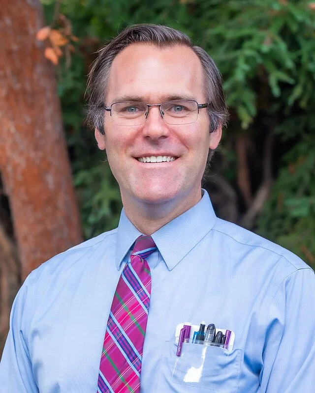 A portrait of a smiling man with glasses, wearing a light blue shirt and a colorful striped tie, standing outdoors with trees in the background.