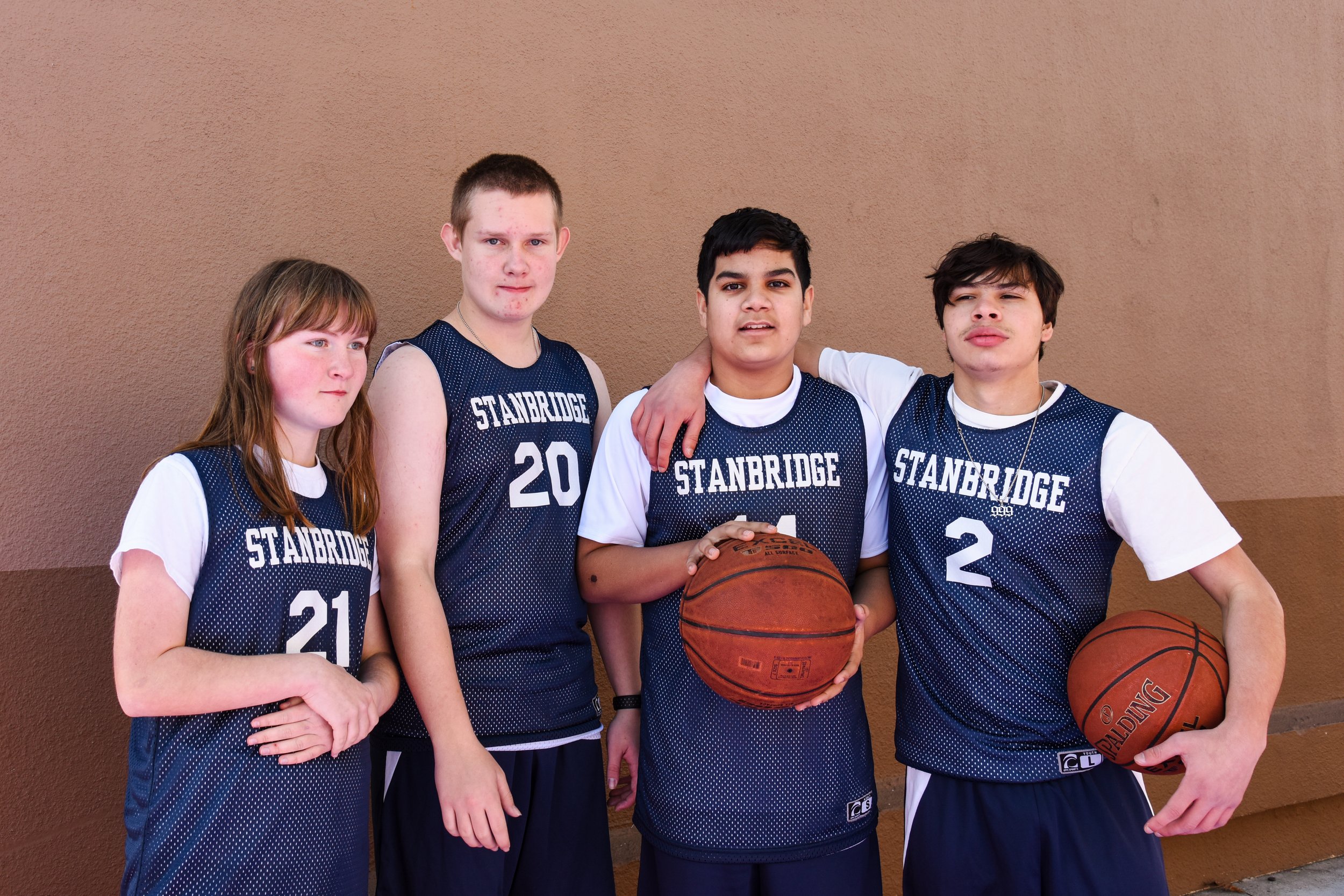 Four young teenagers in basketball uniforms standing against a brown wall, two holding basketballs, with some placing their arms around each other's shoulders.