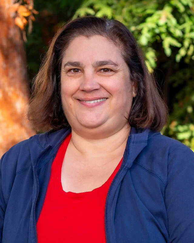 A woman with shoulder-length brown hair smiling outdoors in front of greenery and a tree, wearing a red shirt and a blue jacket.