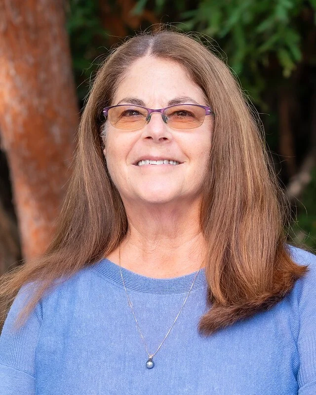 A middle-aged woman with long brown hair, wearing glasses with purple frames, a blue shirt, and a silver necklace with a pearl pendant, standing outdoors with green foliage in the background.