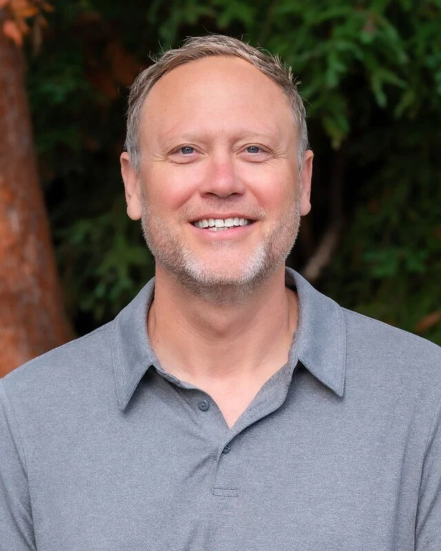 A middle-aged man with short light brown hair, blue eyes, and a beard, smiling in front of green foliage and a tree trunk, wearing a gray collared shirt.
