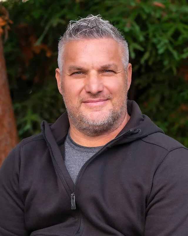 A middle-aged man with short gray hair and a beard, smiling outdoors with green foliage in the background, wearing a black zip-up jacket over a gray shirt.