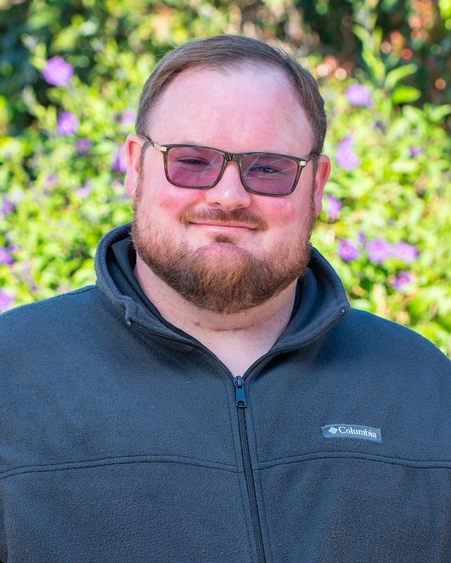 A man with glasses, a beard, and light skin smiling outdoors with purple flowers and green foliage in the background.