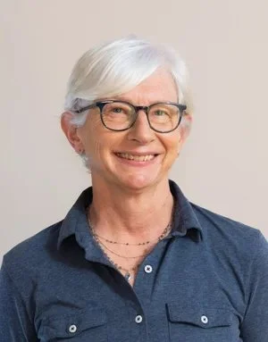 A smiling older woman with white hair, wearing glasses and a dark blue collared shirt, against a neutral background.