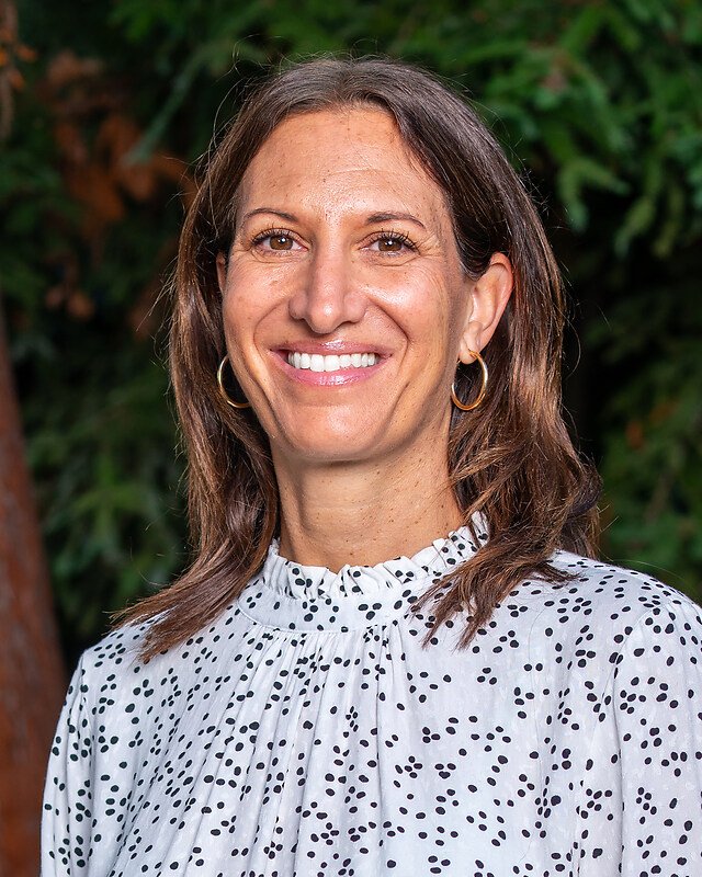 A woman with shoulder-length brown hair, wearing gold hoop earrings and a white blouse with black polka dots, smiling outdoors with greenery in the background.