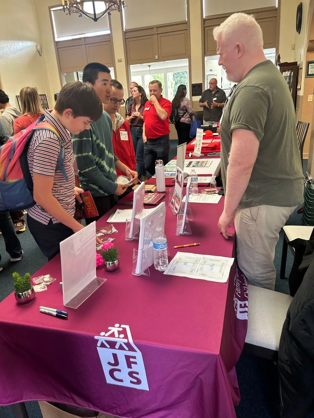 A group of young students and an adult man engaging in conversation at an informational table with pamphlets and signs at an indoor event.