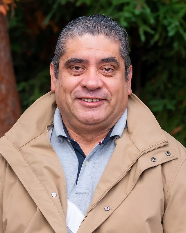 Close-up photo of a middle-aged man with slicked-back gray hair, wearing a beige jacket over a gray polo shirt, standing outdoors with green foliage in the background.