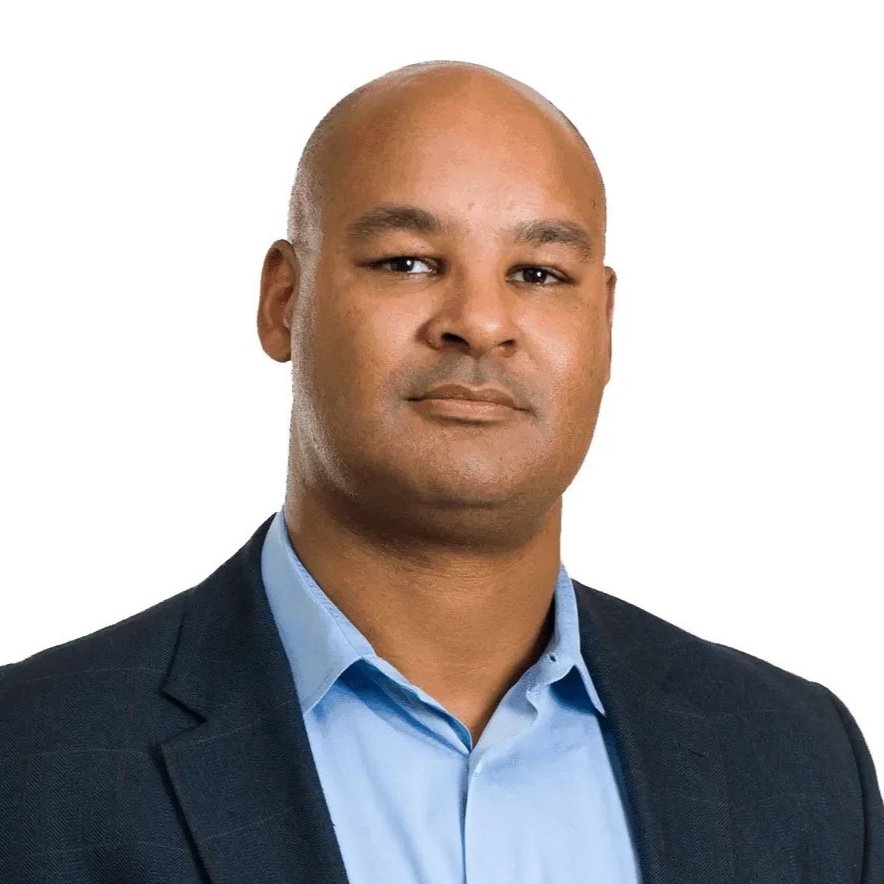 Professional headshot of a bald man in a blue collared shirt and dark blazer against a white background.