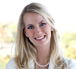 A smiling woman with long blonde hair, wearing pearl earrings and a pearl necklace, outdoors with a blurred natural background.