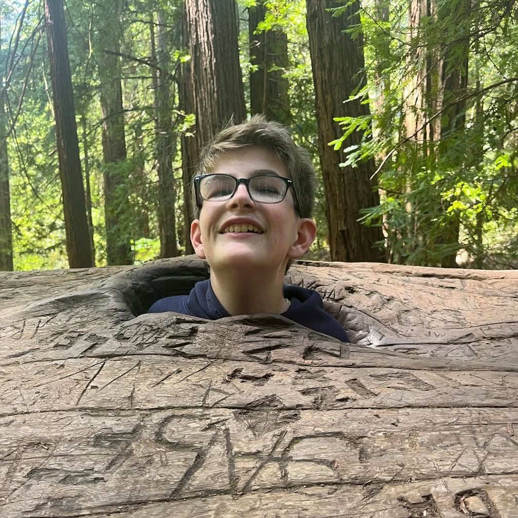 A young person with glasses and short brown hair smiling while leaning on a large fallen tree trunk in a forest with tall trees and green foliage.