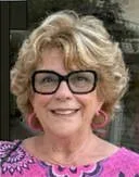 Smiling woman with curly blonde hair wearing glasses and a pink patterned top.