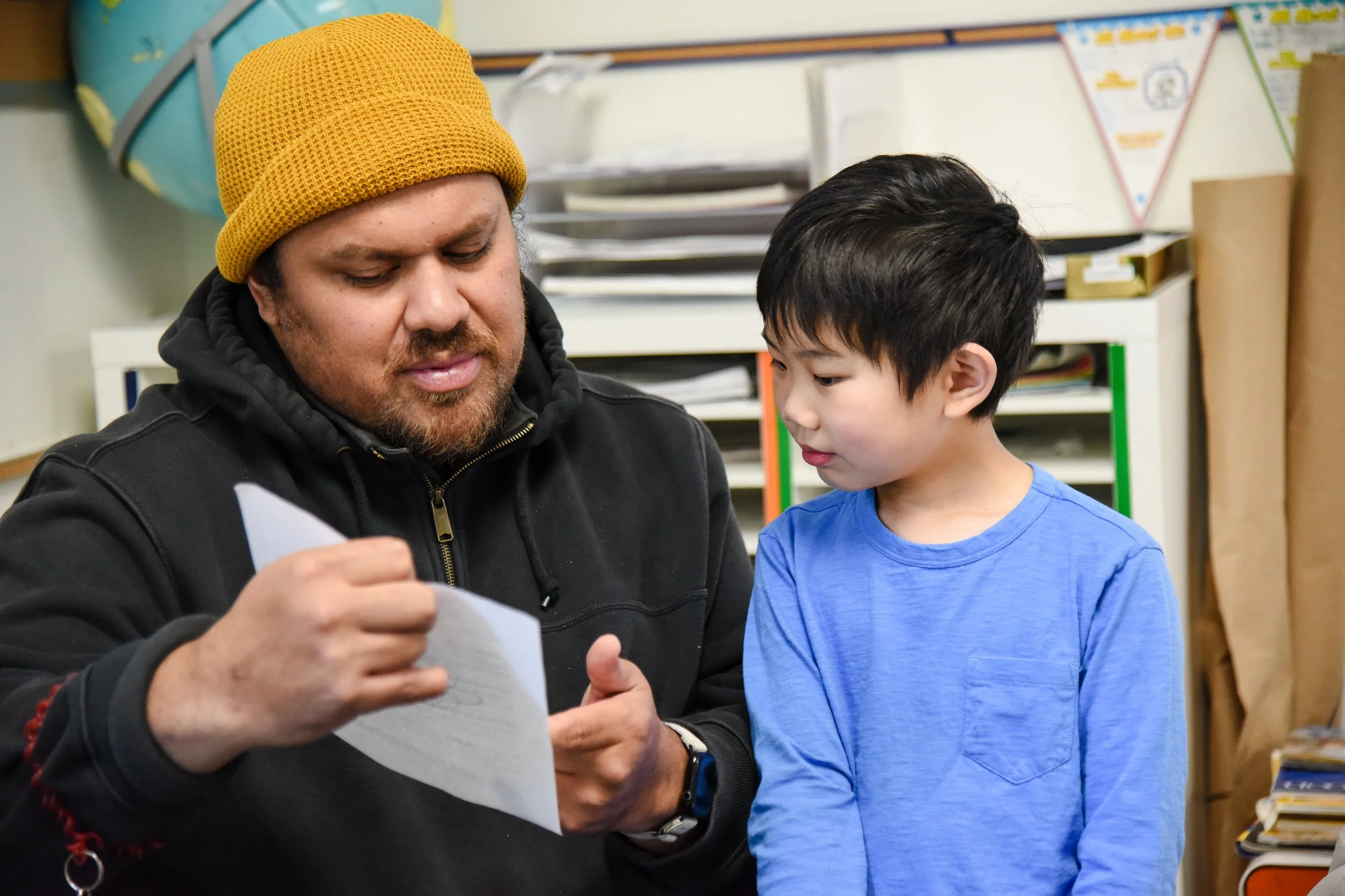 A man and a boy are looking at a piece of paper or a worksheet together in a classroom or library setting.