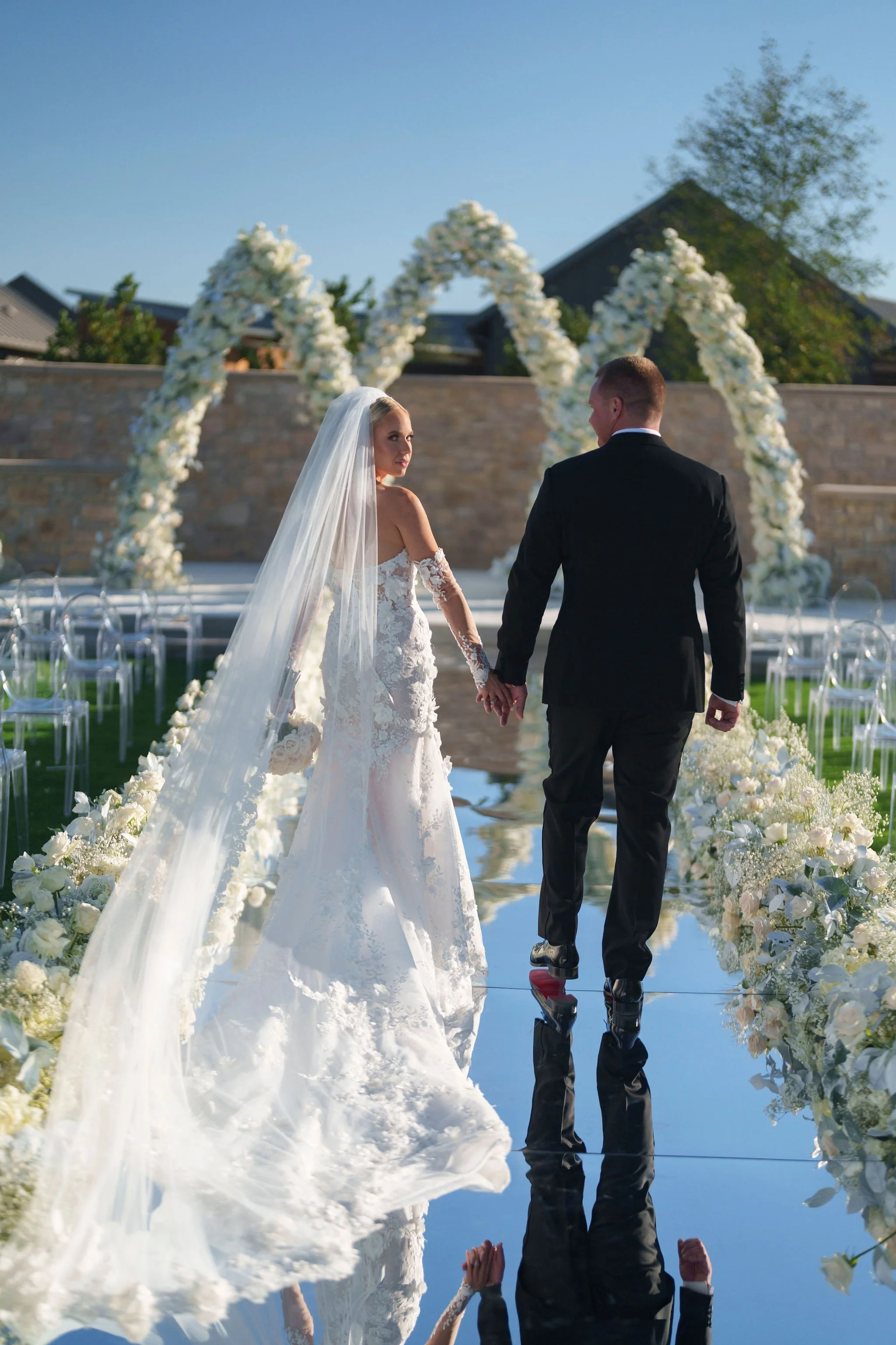 A bride and groom holding hands during their outdoor wedding ceremony, standing under a floral arch with white flowers on a pathway lined with flowers.