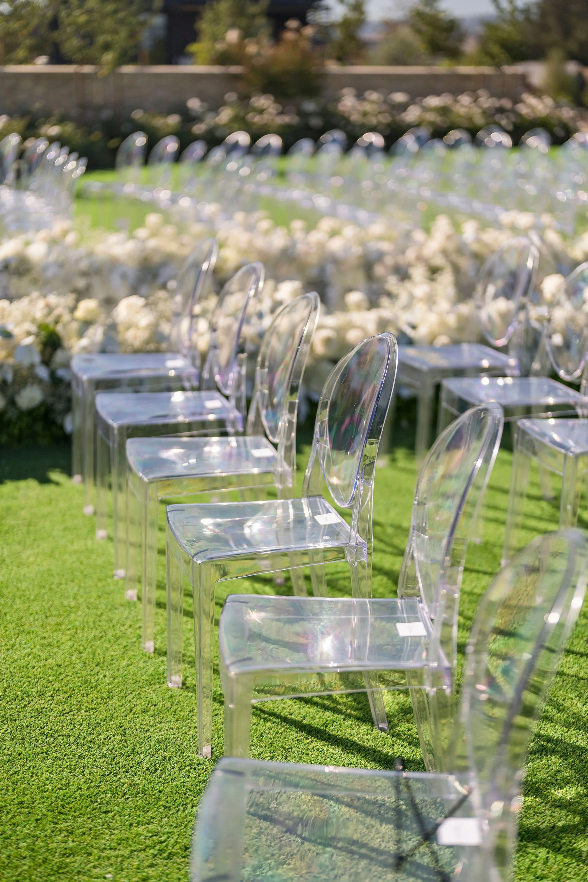 Clear acrylic chairs arranged outdoors on green grass, with white flower arrangements in the background, likely set up for an outdoor wedding ceremony.