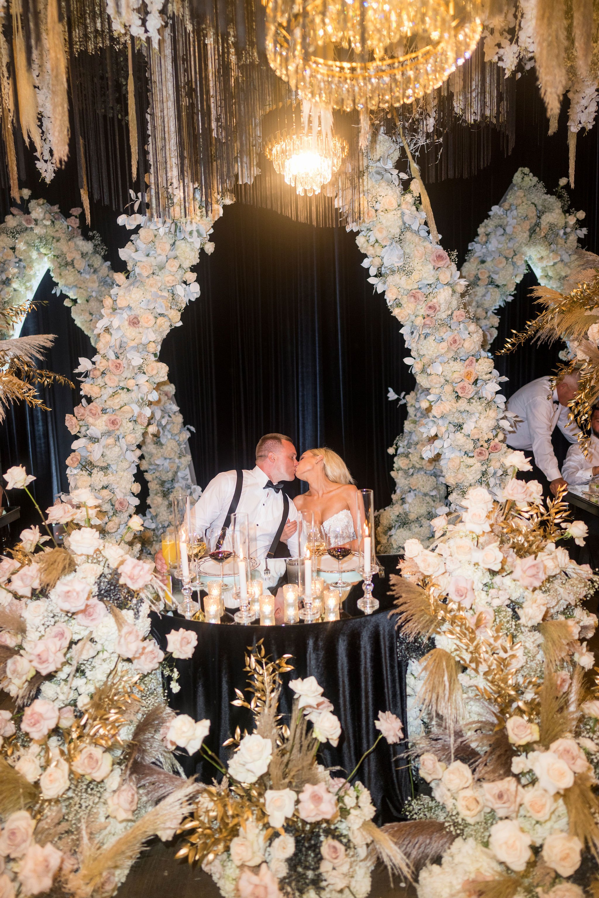A couple in wedding attire kissing in front of a floral arch, decorated with white and light pink roses, gold ornamental leaves, and hanging floral arrangements beneath a large chandelier.