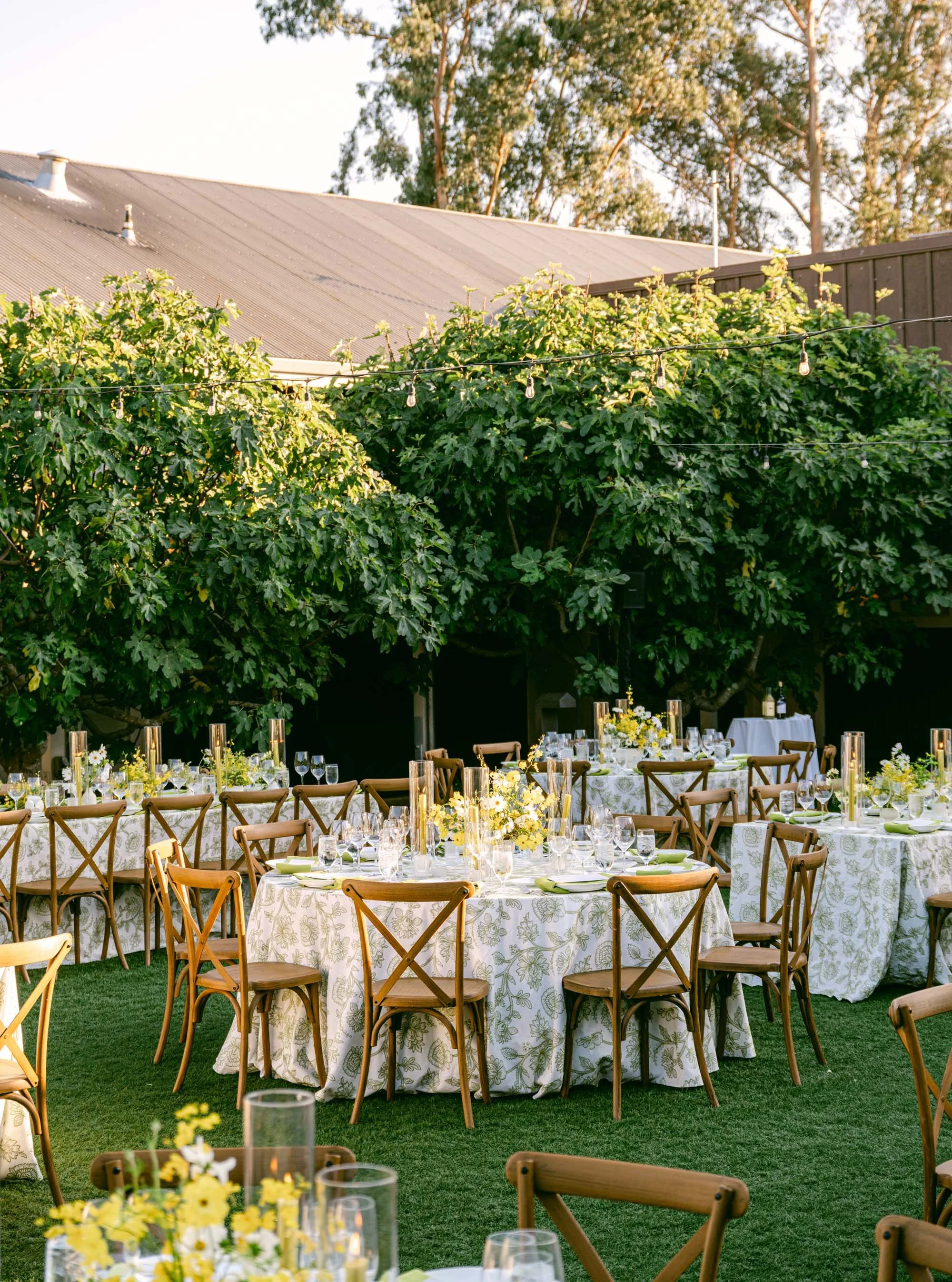 Outdoor dining area with round tables covered with white floral tablecloths, set with glassware, plates, and yellow floral centerpieces. Wooden chairs surround the tables, and string lights are hung overhead. In the background, lush green trees and a building with a metal roof are visible.
