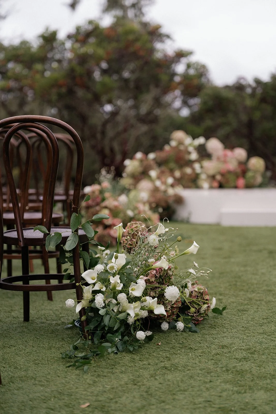 Wooden chairs arranged outdoors with a floral decoration on a grassy surface, including white roses, calla lilies, and other flowers, with trees and bushes in the background.