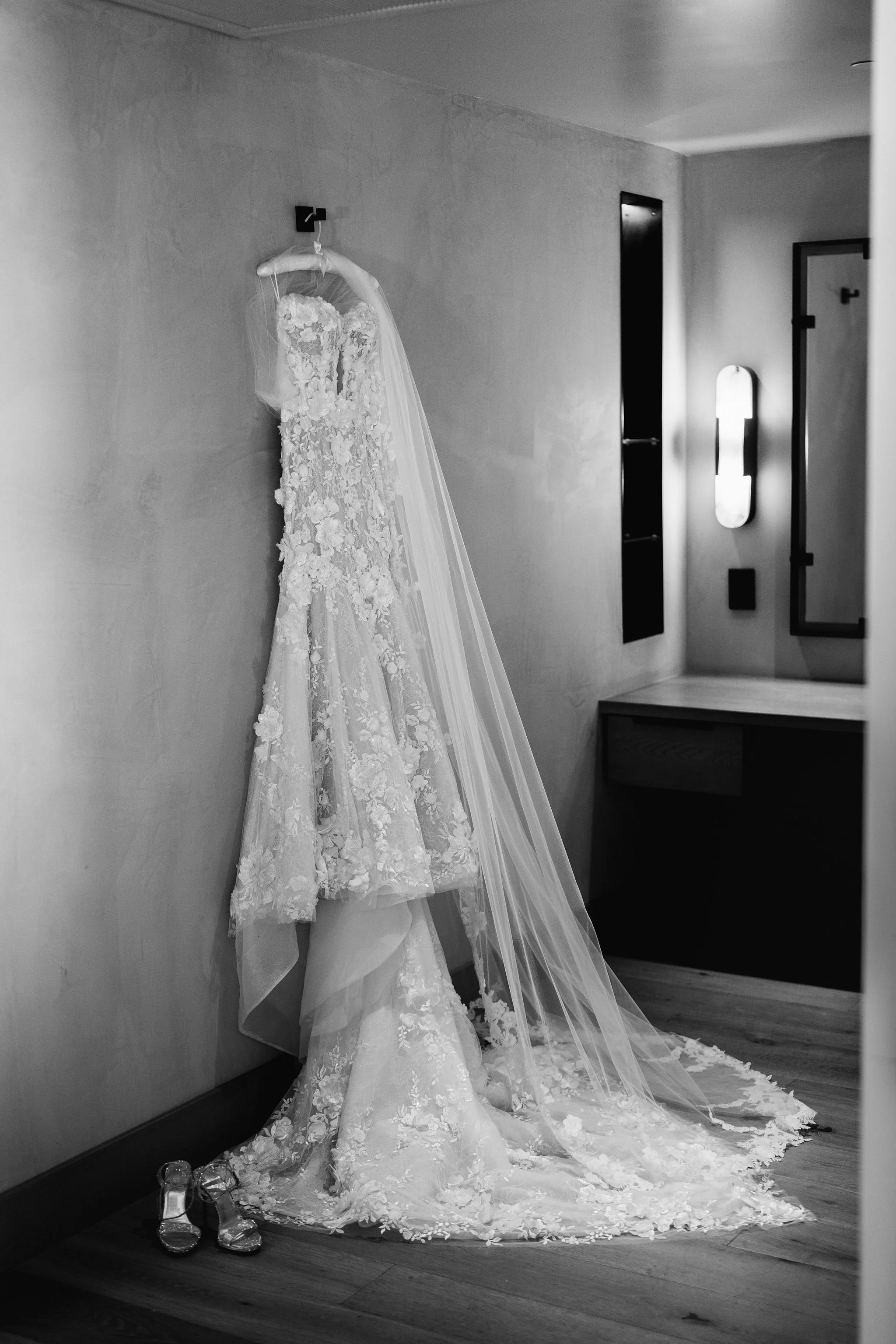 A wedding dress with lace details and a long train hanging on a hanger, paired with a pair of clear, high-heeled shoes placed on the floor next to the dress, in a room with minimal decor and a modern light fixture.