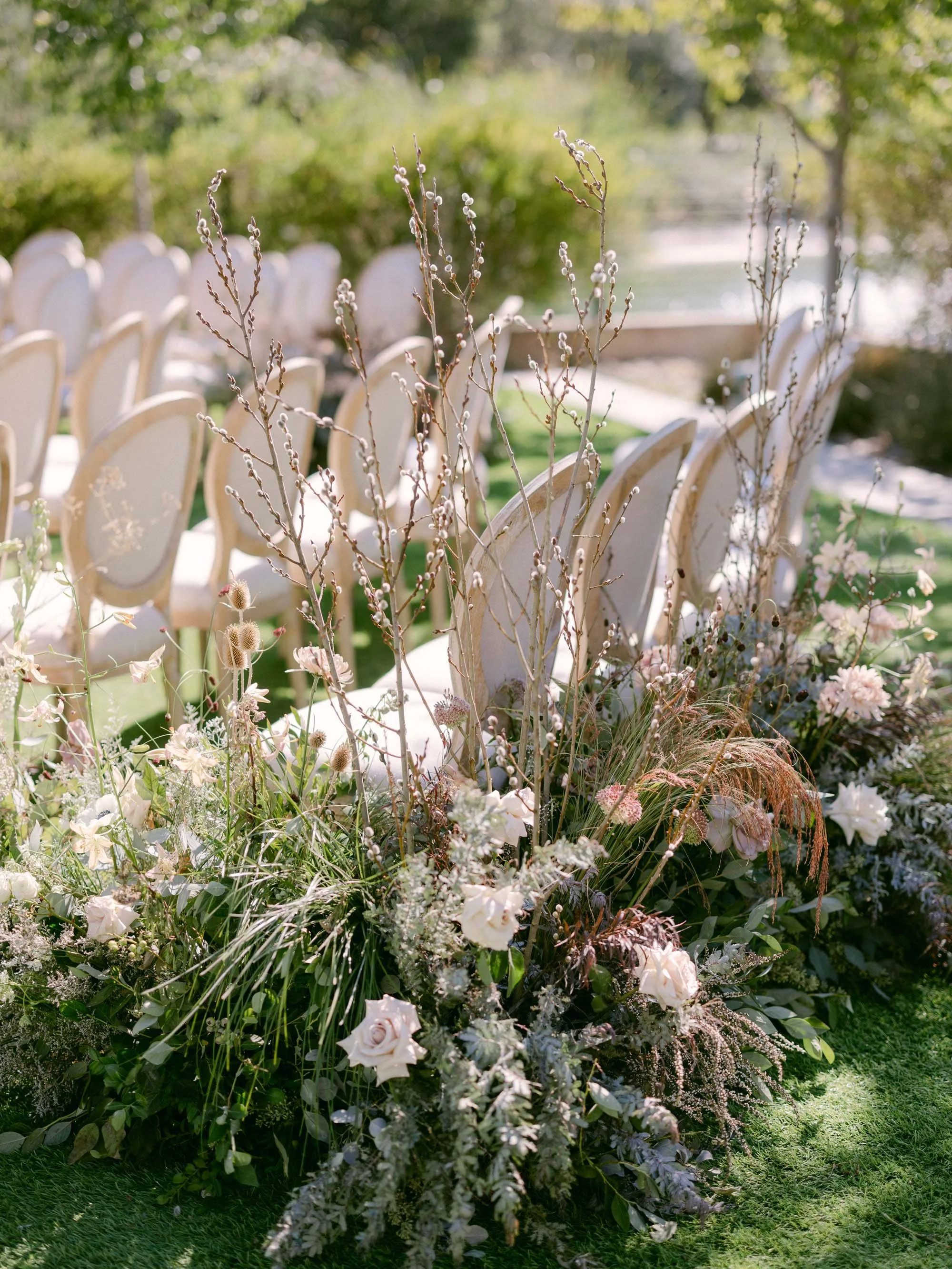 Beautiful outdoor wedding decoration featuring an arrangement of white and blush flowers, greenery, and branches with soft sunlight and a blurred outdoor background.