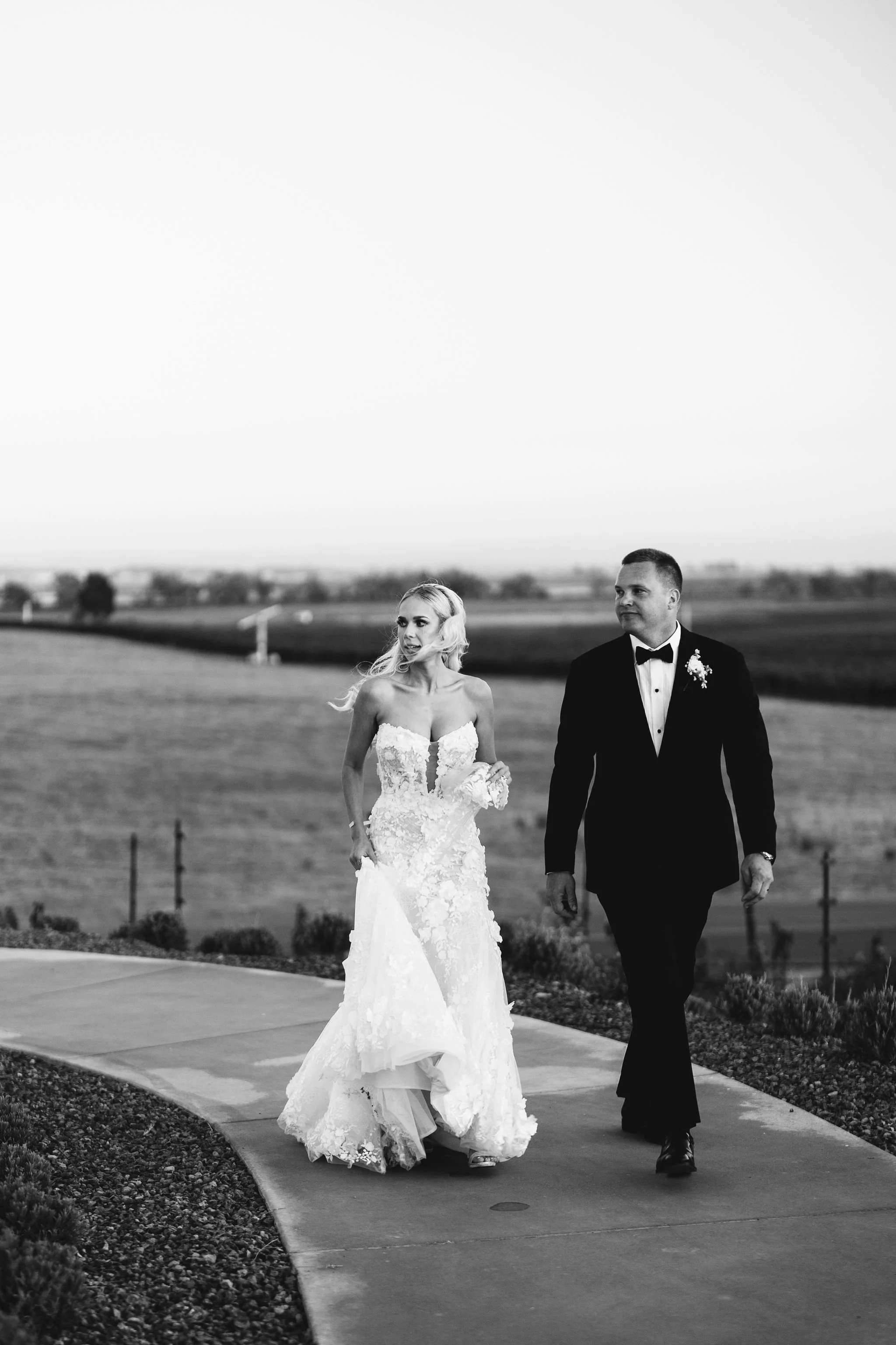 Black and white photo of a bride and groom walking outdoors on a paved path; the bride is in a strapless lace wedding gown holding a bouquet, and the groom is in a tuxedo with a bow tie and boutonniere, walking side by side across a landscaped area w