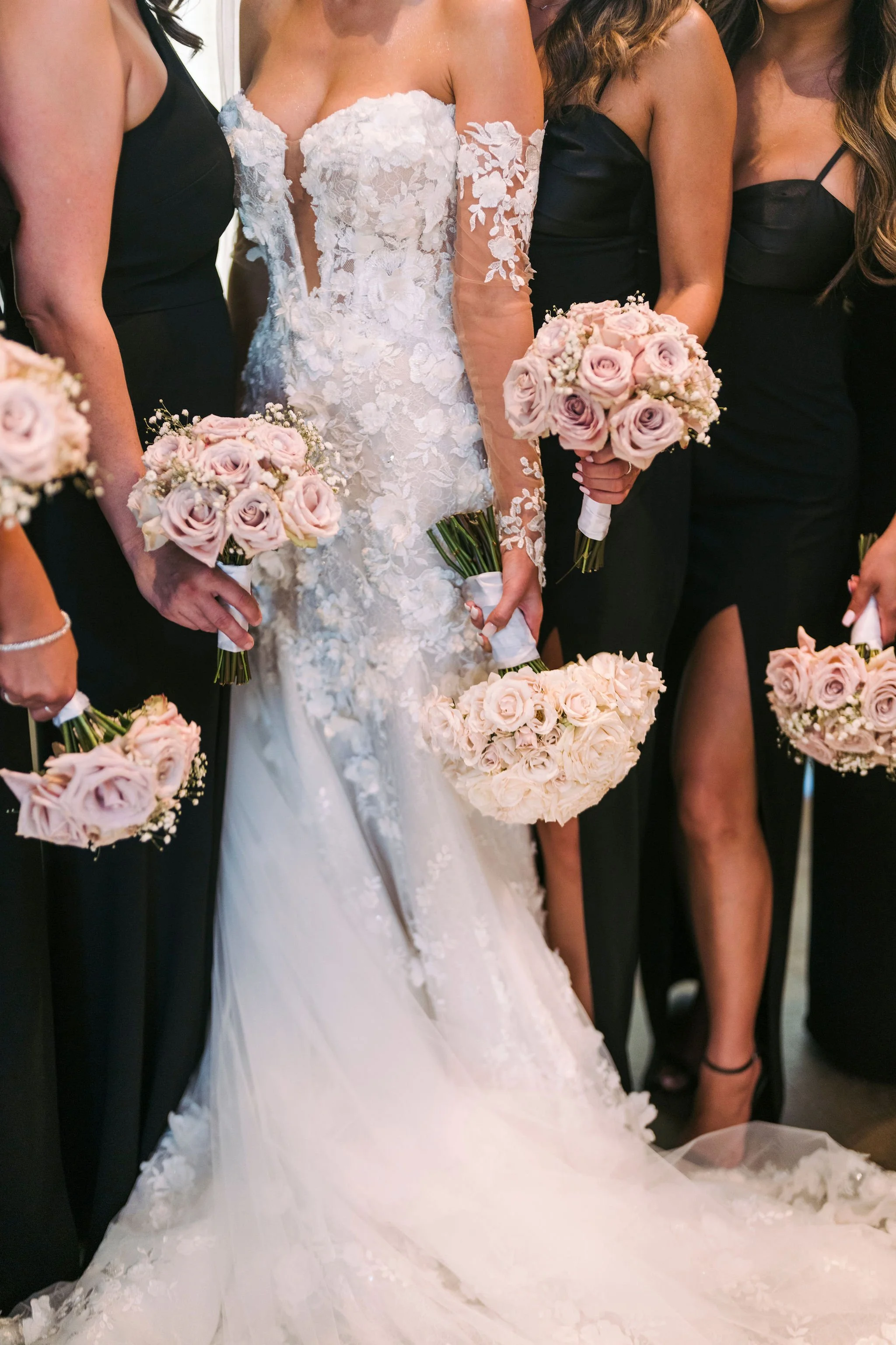 Close-up of women in formal attire holding bouquets, the central figure in a white lace wedding gown, surrounded by bridesmaids in black dresses.