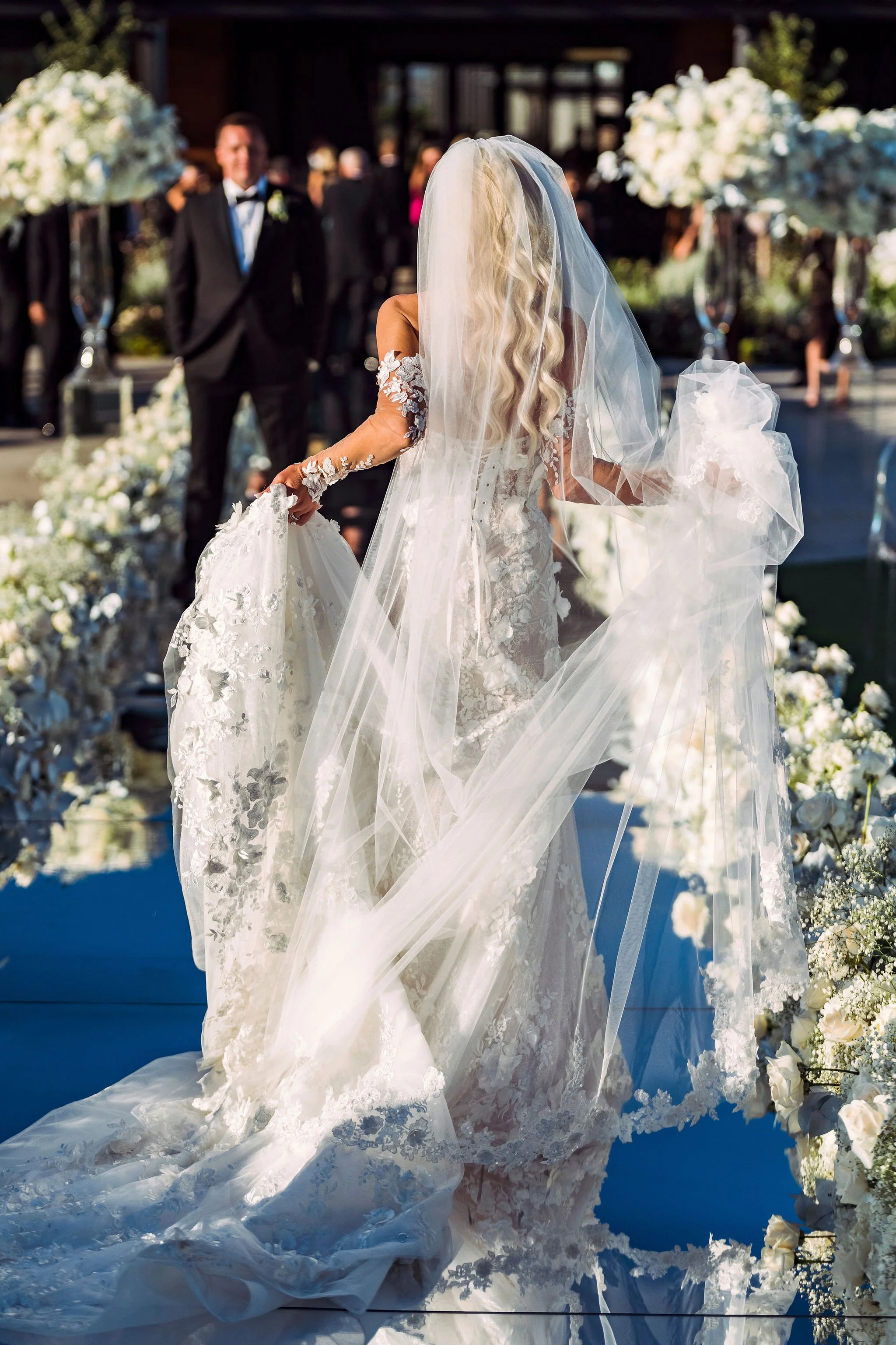 Bride in a white lace wedding dress with floral embroidery, holding her dress with long flowing veil, standing outdoors at a wedding ceremony with floral arrangements and a groom in a black tuxedo in the background.