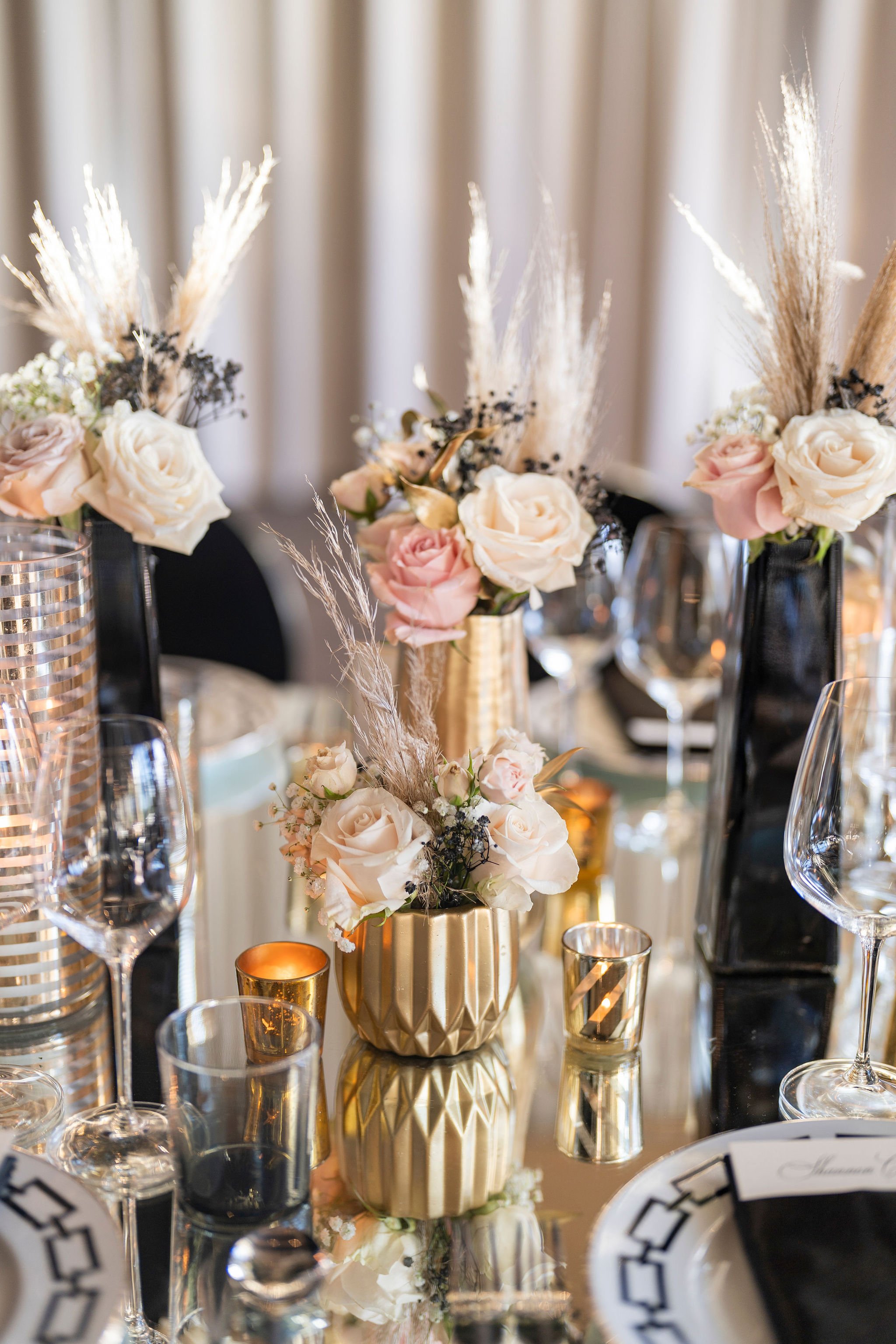 Elegant table centerpiece with white and blush roses, dried plants, and black accents, surrounded by glassware and candles at a formal event.