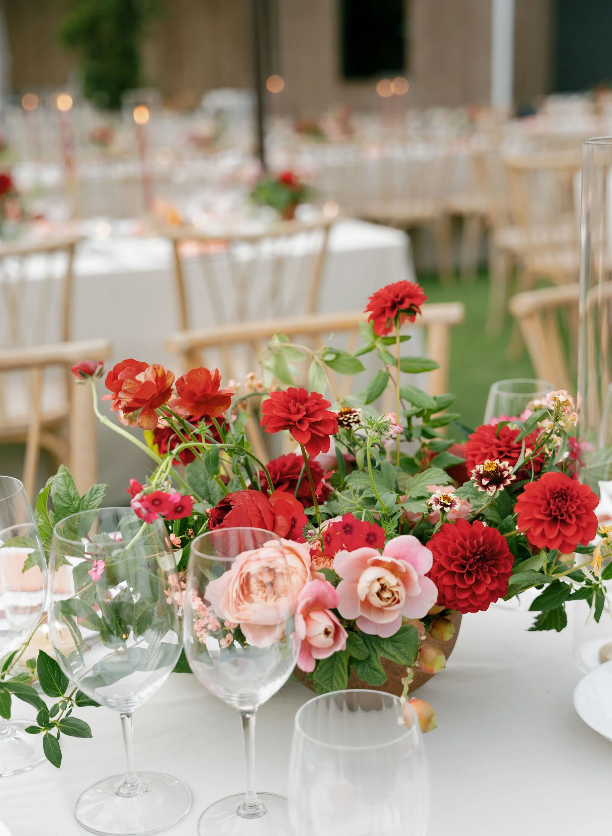 A centerpiece with red, pink, and peach flowers on a table set for an event, with empty wine glasses surrounding it. In the background, there are blurred chairs and tables with similar table settings.